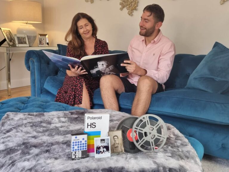 Two people looking and smiling at a large photo album with a baby on the front of it. A mix of cine reels and video tapes are on the table in front of them