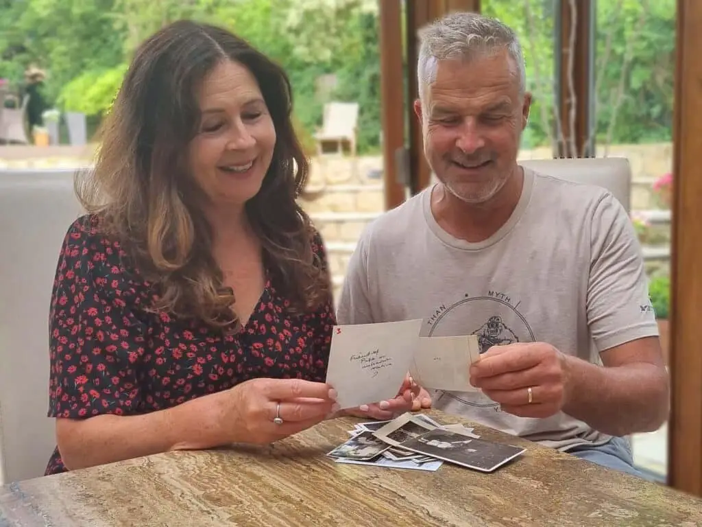 A man and a woman smiling at some photographs laid out on a table with a garden behind them